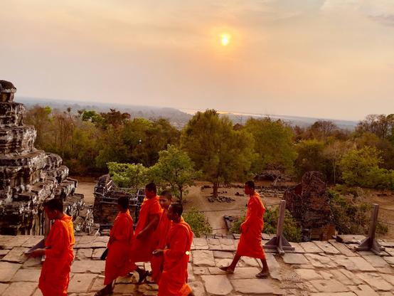A group of teenaged novice monks walk atop the ruins at Phnom Bakheng. The sun is setting the background.