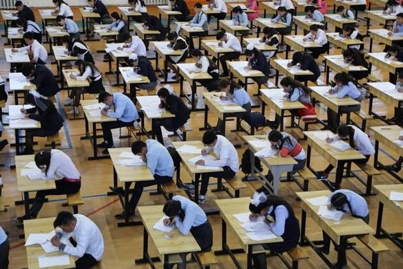 A group of GCSE students sit at light brown desks, all focussing on working. The markings on the floor indicate that it is usually used as a sports hall.