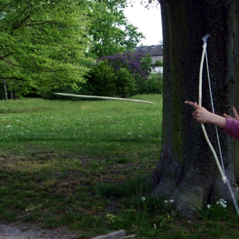 Vor dem grünen Hintergrund einer Wiese  ist ein Pfeil in der Luft zu erkennen. Er wurde gerade abgeschossen, man sieht noch die Kinderhände, die den Bogen führen. Der helle Bogen hebt sich vor einem dunkel berindeten Baum gut ab.