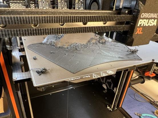 Photo of the printer bed of a Prusa XL 3D printer. A failed print is visible on the printer bed with large filament blobs scattered over the place. The print is partially ripped off the printer bed.