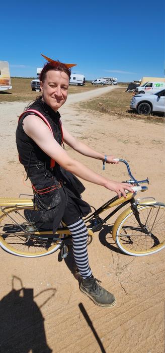 I am sitting on the bike in the dusty Mojave desert. In the background are camper vans. The sun is in my eyes, I am wearing Kamina glasses on my forehead. I have a distressed h.NATO top on, with a skirt, and wearing saddle bags held up by red and white suspenders. I am wearing long stockings, and Doc Marten boots.