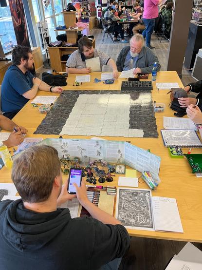 Players sit around the table playing Dungeons and Dragons at Hairy Tarantula. There is a map in the center of the table, and you can see the Dungeon master has several miniatures, papers, and dice hidden behind the Dungeon Master screen.