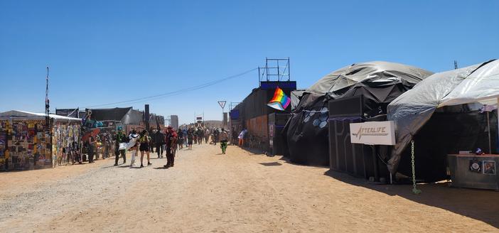 Entering the festival on a Wednesday Afternoon. Along a dirt road is a crowd of futureisticly costumed people. On each side are temporary structures designed to look cyberpunk. From the left is a shed covered in posters. A club named KAIJU, a sign "NPC" a pride flag, and a dome with a sign for "AFTERLIFE".