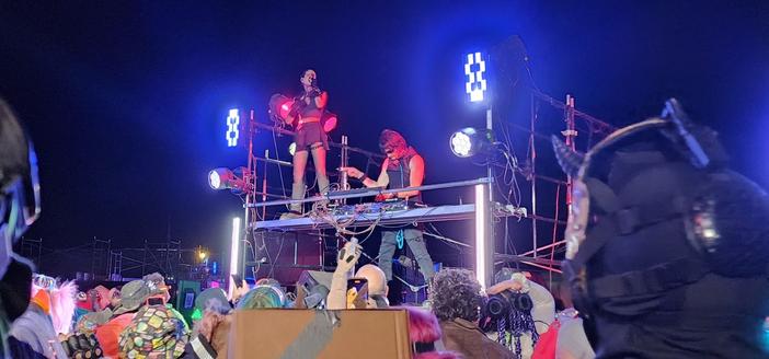 Carah of Shiny Toy Guns, standing on a stage over a crowd at "The Core" in Neotropolis. Jeremy is at the mixer controls.