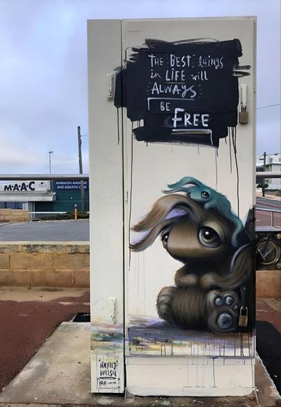 Streetartwall. A mural of two fluffy fantasy creatures has been painted on a beige electricity distribution box near the beach. A small, olive-colored, fluffy creature with very large eyes, curved ears and a broad nose is sitting there with a friend. A smaller green creature is sitting on his head and holding on to him. Above the two of them is a black field with the inscription: 
"The best things in life will always be free". (A low stone wall and the parking lot in front of a maritime clubhouse can be seen in the background. The mural is a popular photo motif for tourists. )