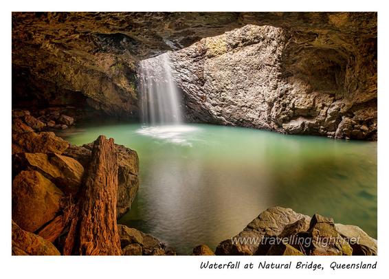 Photo of a small waterfall through the roof of a cave framed by foreground rocks.