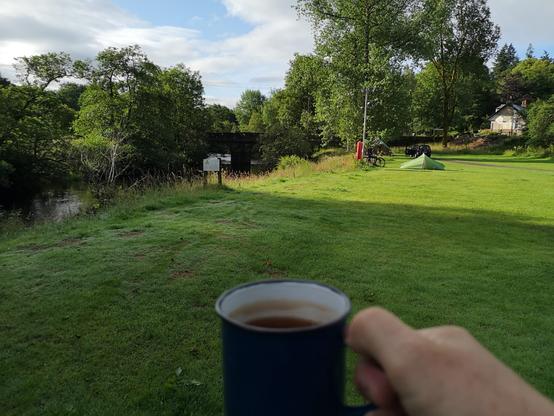 ich halte eine blaue Emailietasse in der Hand und schaue auf die Wiese und den angrenzenden Fluss, wo von derSeite die Sonne leicht rein scheint auf dem Cobbleland Zeltplatz bei Aberfoyle
