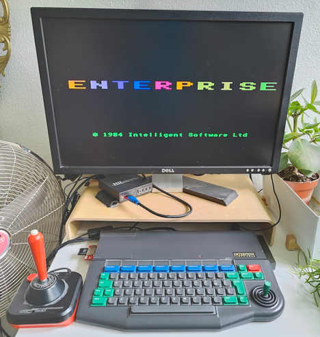 Photo of an Enterprise 64 microcomputer, hooked up to a modern flat panel monitor. Startup screen with color cycling "ENTERPRISE" text visible. Joystick attached. After market SD card reader visible in expansion port.