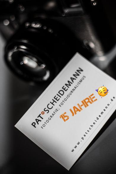 Eine Visitenkarte auf dunklem Hintergrund mit Text und einer Grafik, die auf ein 15-jähriges Jubiläum hinweist, neben einer unscharfen Kamera.

A business card on a dark background with text and a graphic indicating a 15-year anniversary, placed next to a blurry camera.
