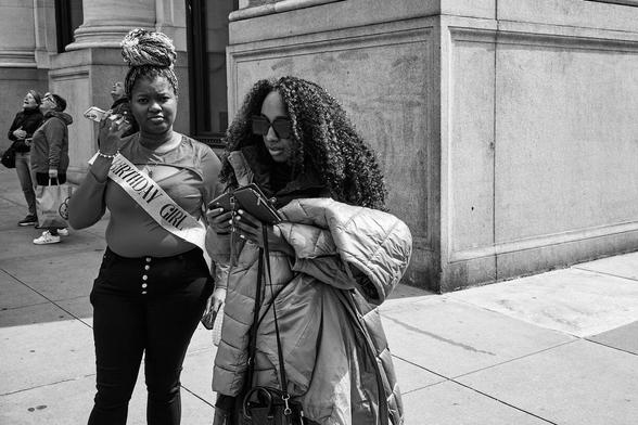 The image is a black-and-white street photograph featuring two women. The woman on the left is wearing a tight-fitting top with the word "FANCY" written across it, pants, and is holding a cell phone in her right hand. She has her hair styled in a bun on top of her head and is looking at the camera with a neutral expression. The woman on the right is wearing large sunglasses, a puffy coat, and a crossbody bag, also looking down at her phone. They are standing near a building with classical architecture; there's a column visible in the background. Both women seem engaged in a common activity, perhaps navigating or scheduling something on their phones.