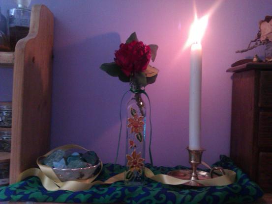 My altar. A small white floral bowl filled with crystals, a floral glass vase with a rose inside tied with a green ribbon, a tall white candle lit in a bronze candle holder, all on a piece of green and blue cloth and draped in yellow ribbon.