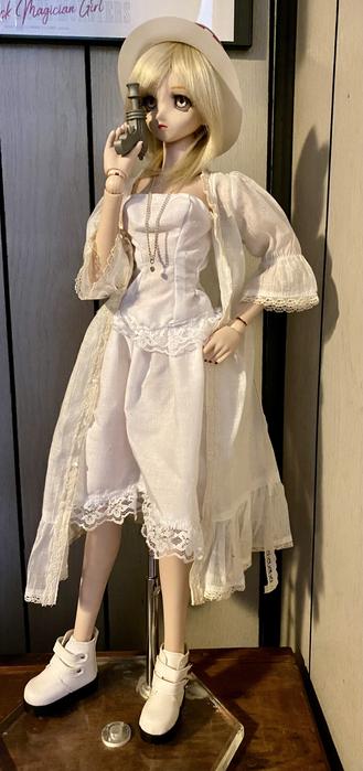 Doll in a white vintage-style dress and hat, holding a blaster near her face, standing on a wooden surface.