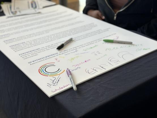 A large poster with the IPSE day proclamation. It has three different colored Sharpie pens sitting on it, and has a number of signatures in different colors on the bottom, with space for more.