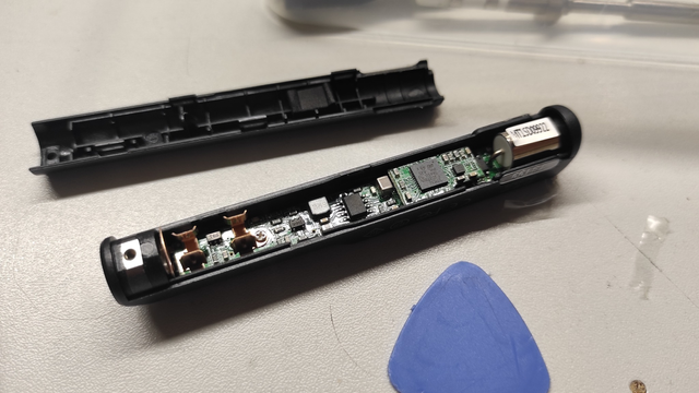 A TS-100 soldering iron case lying open on the table, revealing the printed circuit board inside. The other part of the case and a plastic prying tool for opening cases are visible nearby.
