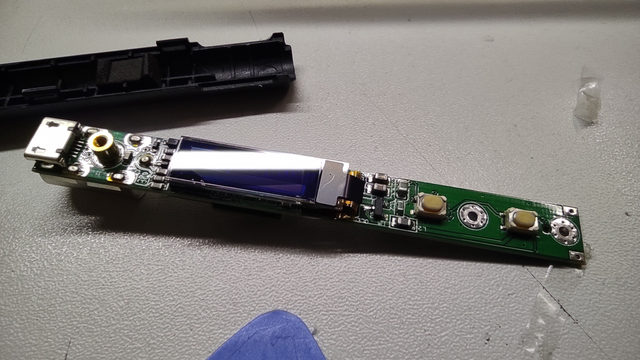 A TS-100 soldering iron PCB lying on the table, removed from both halves of the case. One part of the case and a plastic prying tool for opening cases are visible nearby.