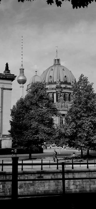 Black and white photo of the  cathedral and the TV tower in Berlin,  Germany. In front of the cathedral are two trees with people walking. Immediately in front is the riverside railings. Taken from across the river.