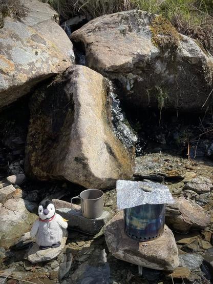 A stove inside a windshield, a mug, and Penguin in a stream next to a mini waterfall.