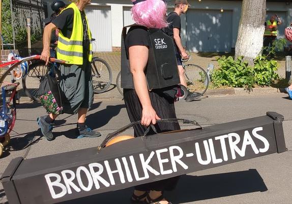 Person mit pinken Haaren und schwarzer Kleidung trägt einen länglichen schwarzen Kasten mit Griffen und Aufschrift 'Brorhilker Ultras'. Sie trägt eine imitierte Schutzweste aus Pappe, schwarz, mit der Aufschrift 'SEK Grunewald'.