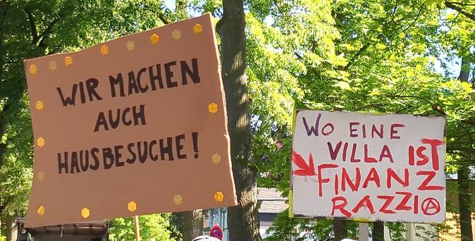 Zwei Demoschilder vor sonnenbeschienenen Bäumen. 

"Wir machen auch Hausbesuche", "Wo eine Villa ist Finanzrazzia"