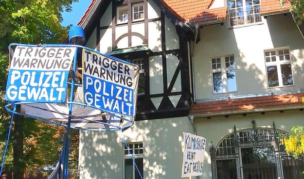 Vor einem großen Haus mit Fachwerk verschiedene Demoschilder. 

Weiß-blau: "Triggerwarnung Polizeigewalt" und "Klimaschutz heißt Eat The Rich"