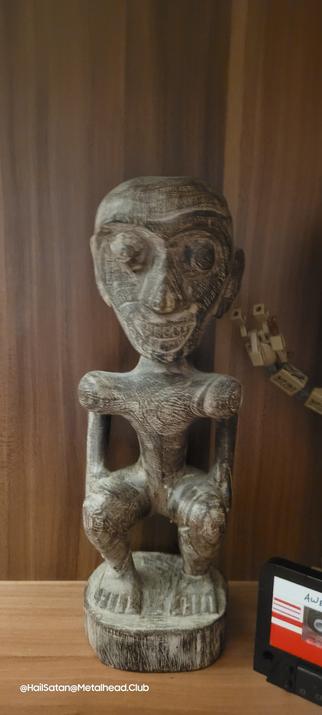 Afrikanische Kunst. Eine Schnitzerei. Es zeigt einen Mann in der Stehhocke, die Hände auf den Oberschenkeln mit weit aufgerissenen Augen und breiten Grinsen.