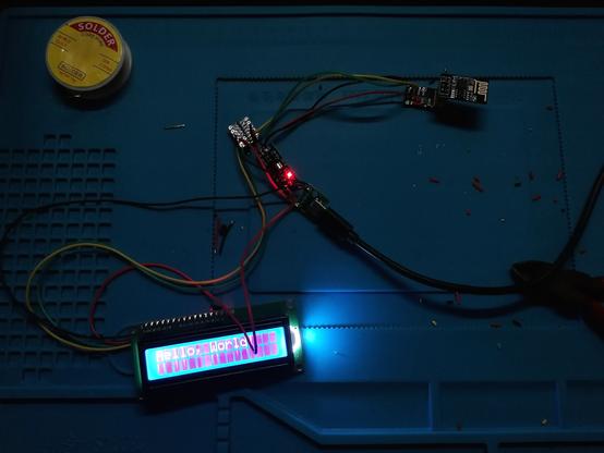 A very dark picture of a bright shining LCD display saying "Hello, World!". Many wires connect the LCD with a level shifter a ESP01 is connected to from the other side and a step-down module with USB. Beside there is soldering wire and a pincers. All is laying on a heat resistent soldering pad