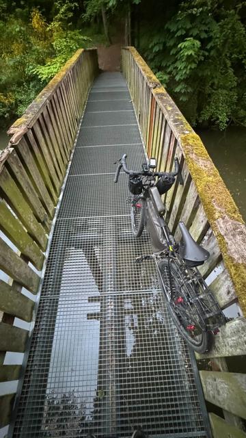 Fahrrad steht auf einer Brücke mit Gitterboden und darunter sieht man den Fluss. Das Geländer der Brücke ist aus Holz ￼