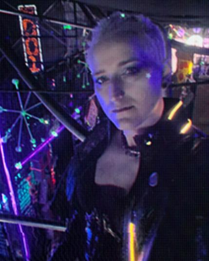 Photo of a person with blonde hair standing on a catwalk above a cyberpunk city street with glowing neon lights. They are wearing a leather jacket with glowing fixtures around the neck.