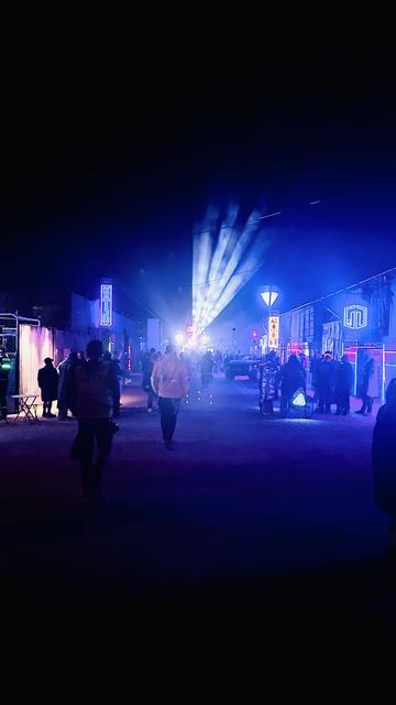 Photograph of the Neotropolis festival. The photo shows a cyberpunk city street built out of metal sheeting and scaffolding. There are neon lights with the festival logo on the right. On the left a neon sign reads “Kaiju Nightclub”. At the end of the street there is a stage with a very large neon sign that reads “the core”. The street is made of sand and there is dust in the air. A few dozen people in cyberpunk outfits wonder up and down.