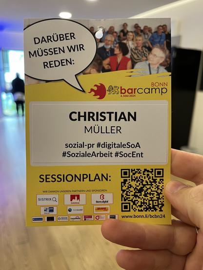 Alternativtext: Ein Foto einer Hand, die eine Namenskarte hält bei einem Event. Die Karte zeigt "Christian Müller" und den Namen "Bonn Barcamp" mit dem Datum 4. Mai 2024. Oben auf der Karte ist ein Bild von Menschen bei einer Konferenz. Die Karte enthält auch die Hashtags #sozialpr #digitaleSoA #SozialeArbeit #SocEnt und einen QR-Code unten rechts, sowie Logos von Sponsoren und Partnern.