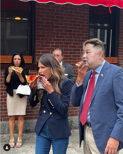 A badly photoshopped Kim Jong Un's head on top of a person wearing a suit eating pizza next to Kristi Noem also eating pizza standing on a sidewalk outside a brick building