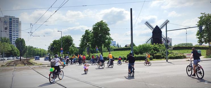 Die Radfahrenden der Kidical Mass Marzahn-Hellersdorf biegen von der Landsberger Allee in die Allee der Kosmonauten ab. Es sind unter den Radfahrenden auch viele Kinder, einige haben die Räder mit Luftballons geschmückt. Im Hintergrund ist die Blockwindmühle in Alt-Marzahn zu sehen. Weiterhin sieht man die Tramschienen und ein großes Wohngebäude links im Bild.