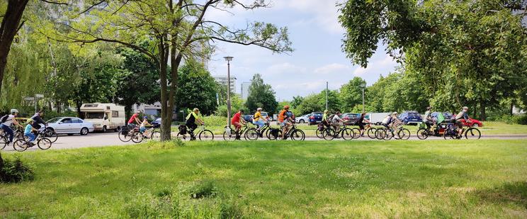Die Radfahrenden der Kidical Mass Marzahn-Hellersdorf fahrenneben einem sommerlich-frühlingshaften Grünstreifen mit ein paar Bäumen auf einer Straße. Das Foto ist von der Seite aufgenommen. Im Hintergrund befindet sich ein Parkplatz mit einigen Autos darauf. Unter den Radfahrenden sind Kinder und einige der Teilnehmenden haben High-Visibility oder auch Warn-Westen an. Einer der Radfahrenden ist ein Polizist der Fahrradstaffel.