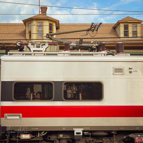 A square cropped photo of the side of a train car. In the window of the car, we can see a woman seated on the train. She is wearing headphones and staring off into the distance. We can also see through the window on the opposite side of the train, to the people standing on the platform on the other side.