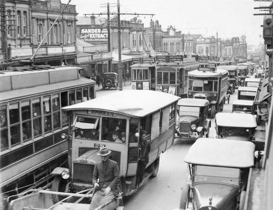 Black-and-white photo: Traffic on Upper Symonds Street, Auckland Central. 1920s. Description: Cars, buses and trams in a traffic jam across from the premises of William Gray, watchmaker, William Wallace, hairdresser, and Watney Sibun and Son on Upper Symonds Street. Citation: Auckland Libraries Heritage Collections 589-0058. https://kura.aucklandlibraries.govt.nz/digital/collection/photos/id/320165