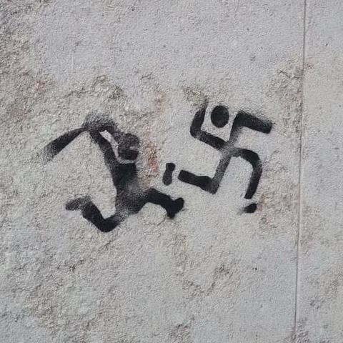 A black ink stencilled graffiti image on a grey stone wall. There is a figure of a running person with mask on wielding a bat at another figure of a person running away. That figure is made up of a swastika at an angle that has a round circle for a head and small shoes on the bottom two limbs.