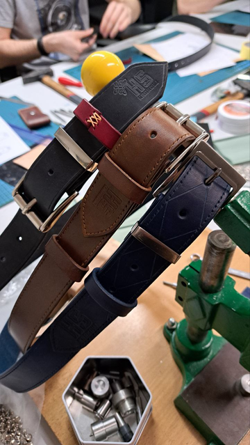 Three leather straps hanging on a handle, one black with a red leather slot stitched with light brown thread, the second brown, and the third navy blue. Each of them has the Hackerspace Kraków logo embossed on the leather, more or less prominently displayed. Tools are visible on the table, and in the background, someone is working on creating their strap.