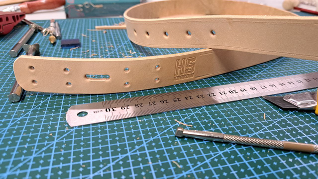 A strap made of light, raw, uncolored leather lying on a cutting mat. The Hackerspace Kraków logo is embossed on it, and next to it lies a metal ruler and leatherworking tools.