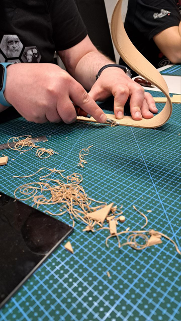 The photo shows the hands of a person working on leather, specifically performing edge beveling - a process of cutting the leather edge at a 45-degree angle to ensure a smooth transition for easier edge polishing later on. Visible on the table are leather scraps and shavings resulting from this process.
