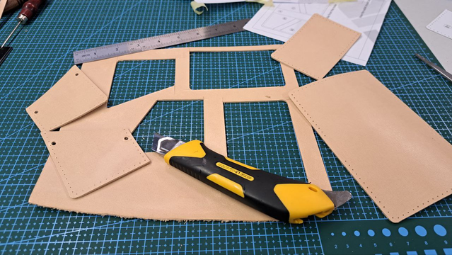 The photo shows a sheet of leather from which rectangular pieces have been cut, likely for a wallet or pouch. On the sheet, placed on a cutting mat, lies a wallpaper knife. In the background, there is a piece of paper with a template or cutting scheme.