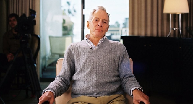 Robert Dust, sentado en un sillón blanco, mirando a cámara, en mitad de un salón de habitación de hotel con la ventana al fondo, mira a cámara, antes de una de las entrevistas del documental.