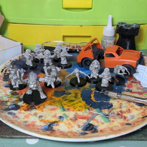 A collection of 12 unpainted metal 28mm scale miniatures to be used as a Genstealer Cult army and an orange toy car that has bee cut in half. These are sitting on a paint splattered plate on a messy desk.
