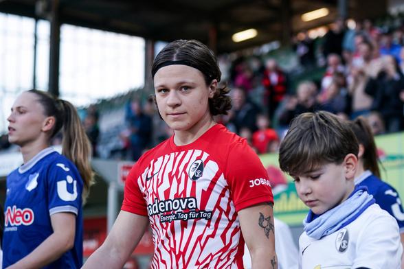 Ally Gudorf im rot-weißen Trikot des SC Freiburg mit einem Einlaufkind an der Hand. Gudorf schaut genau in die Kamera. Im Hintergrund ist die Tribüne mit Fans erkennbar.