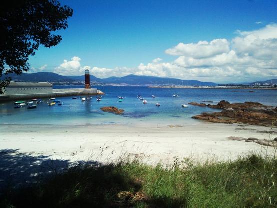 Photo of a small beach. Just before the beach some grass and a tree can be seen. Right after the grass the beach starts with some sand and a calm sea from an estuary with an intense blue color. In the background some mountains can be seen with some villages on their base.
On the left side there is a small pier that ends with a lighthouse.
There are some clouds in the sky.