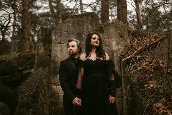 In the image, a couple stands in a moody, natural setting that emphasizes a sense of both elegance and solitude. The bride wears an alluring black gown with sheer, flowing details on the sleeves, contributing to a dramatic and somewhat gothic aesthetic. Her makeup is bold, featuring dark lipstick that complements her full-length dress. She stands confidently beside her partner, looking into the distance with a powerful, contemplative expression. The groom, slightly in the foreground, is dressed in a sleek black suit, his hair styled neatly, and he gazes off to the side with a serious demeanor. They are surrounded by the rugged textures of ancient, crumbling stone structures and bare trees, enhancing the dark, timeless theme of the setting. The overall atmosphere is one of stark beauty and an intimate connection shared in a secluded, almost mystical landscape.