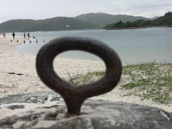 Blick durch einen Eisenring am Ufer der White Sands of Morar, auf eben diesen weißen Sandstrand. Rechts im Bild sieht man eine Flussmündung, die ins Meer geht und auf der linken Bildseite, den weißen Sandstrand, der aus dem Film "lokal Hero" bekannt ist