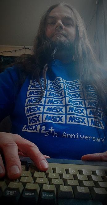 FiXato, a bearded man with long hair, wearing an MSX2+ 25th Anniversary t-shirt, with his hands resting on the keyboard of a Philips NMS8250 MSX2 homecomputer.
