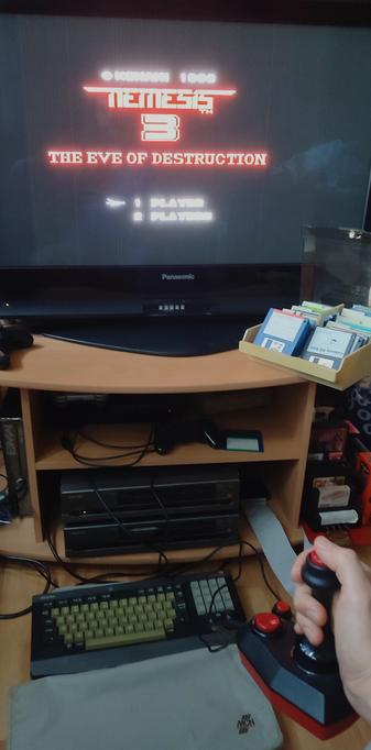 Title screen of Nemesis 3: The Eve of Destruction on an old widescreen LCD TV. In front of the TV is an open diskette box with 3.5 inch diskettes, and below it are two Philips NMS8250 MSX2 homecomputers, with the top one powered on. On the floor in front of it is the keyboard, and an MCN keyboard cover.
A hand holding a two-button "The Arcade Turbo" joystick can be seen in the bottom right corner, and just above it a (not connected) slot expander with a Philips Music Module, a Sunrise CF reader, a Panasonic FM-PAC, and a 1MB Memory Mapper.