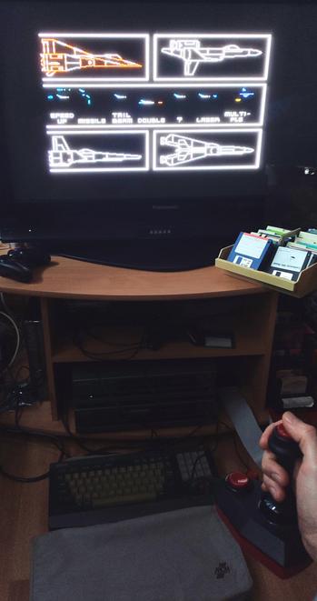 Fighter Jet configuration screen of Nemesis 3: The Eve of Destruction on an old widescreen LCD TV. In front of the TV is an open diskette box with 3.5 inch diskettes, and below it are two Philips NMS8250 MSX2 homecomputers, with the top one powered on. On the floor in front of it is the keyboard, and an MCN keyboard cover.
A hand holding a two-button "The Arcade Turbo" joystick can be seen in the bottom right corner, and just above it a (not connected) slot expander with a Philips Music Module, a Sunrise CF reader, a Panasonic FM-PAC, and a 1MB Memory Mapper.