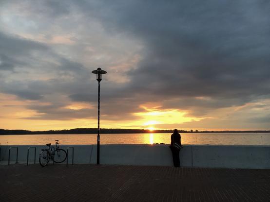 Sonnenuntergang aus einem Wolkenfeld heraus über der Förde, spiegelt sich lang im Wasser. Im Vordergrund an einer Mauer ein Mensch als Schattenriss, eine Lampe und daneben ein Rad an Halter angelehnt.